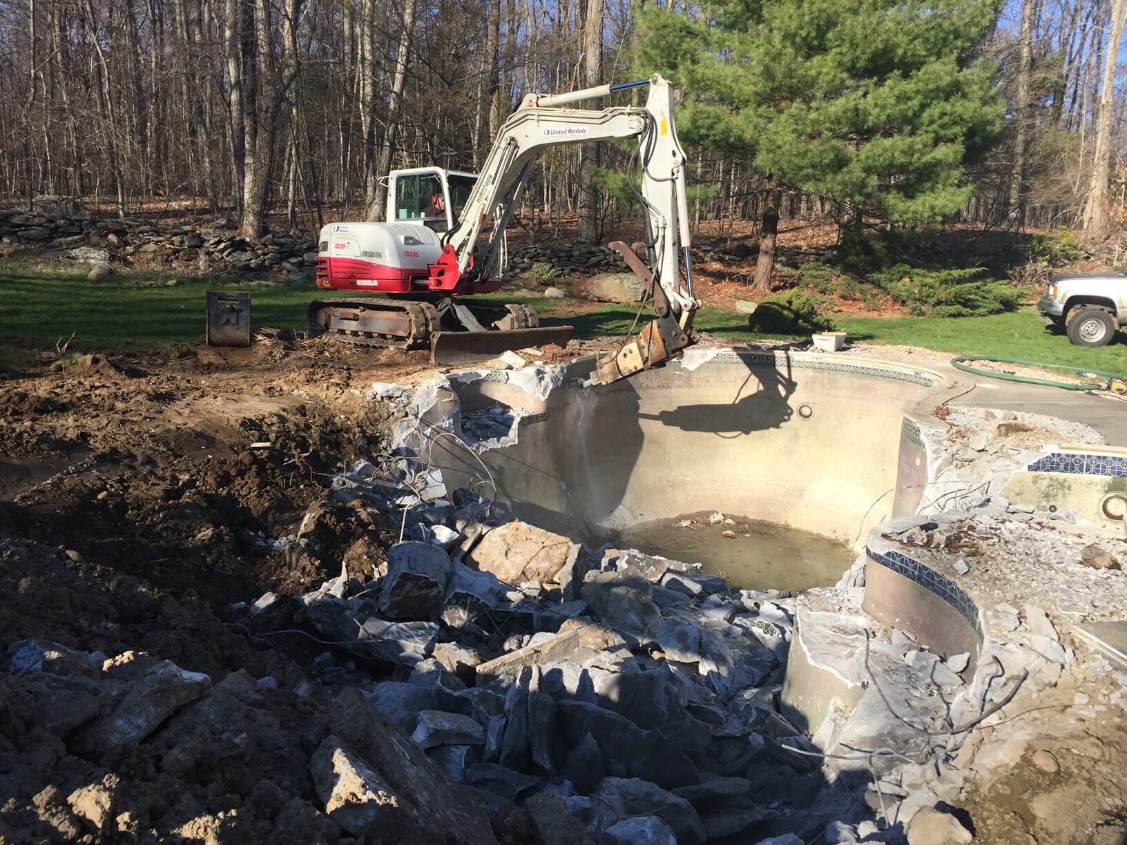 An excavator with a hydraulic breaker attachment is used to break up gunite pool.